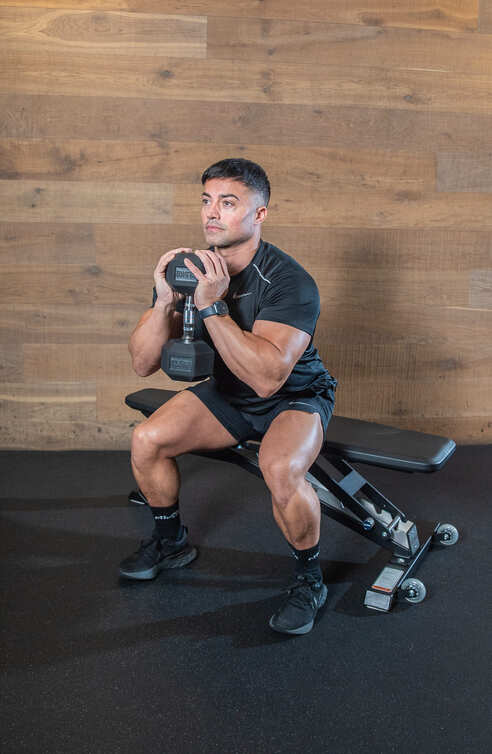 holding a weight with both hands at chest height, man bends his knees to demonstrate goblet squat