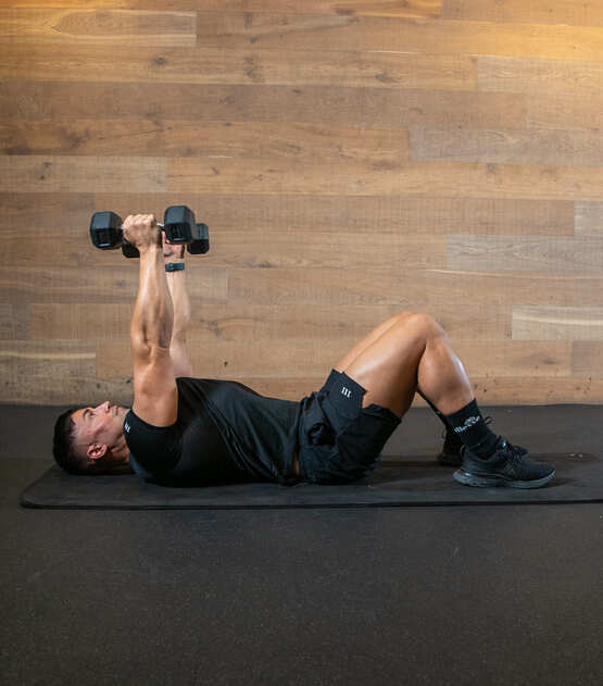 Part of the best full-body dumbbell workout: man demonstrated rolling dumbbell extension; laying on floor with knees bent, he lifts two dumbbells overhead