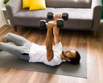A man lying on an exercise mat in his living room, performing dumbbell presses