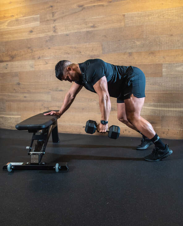 Part of the best full-body dumbbell workout: alternating bent over dumbbell row demonstration; man bends forwards with one hand on bench, the other holding a dumbbell