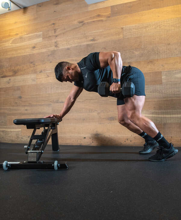 Part of the best full-body dumbbell workout: alternating bent over dumbbell row demonstration; man bends forwards with one hand on bench, the other rows a dumbbell up and down