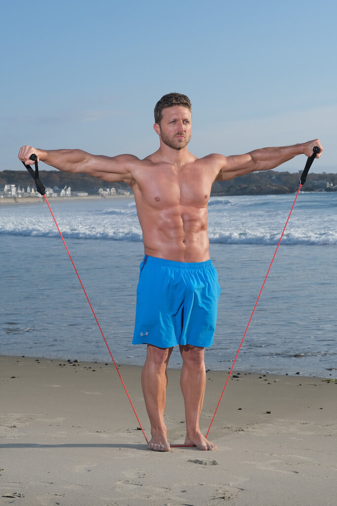 man demonstrates resistance band lateral raise. Holding a resistance band in each hand and under his feet, he raises each arm under level with his shoulders before lowering