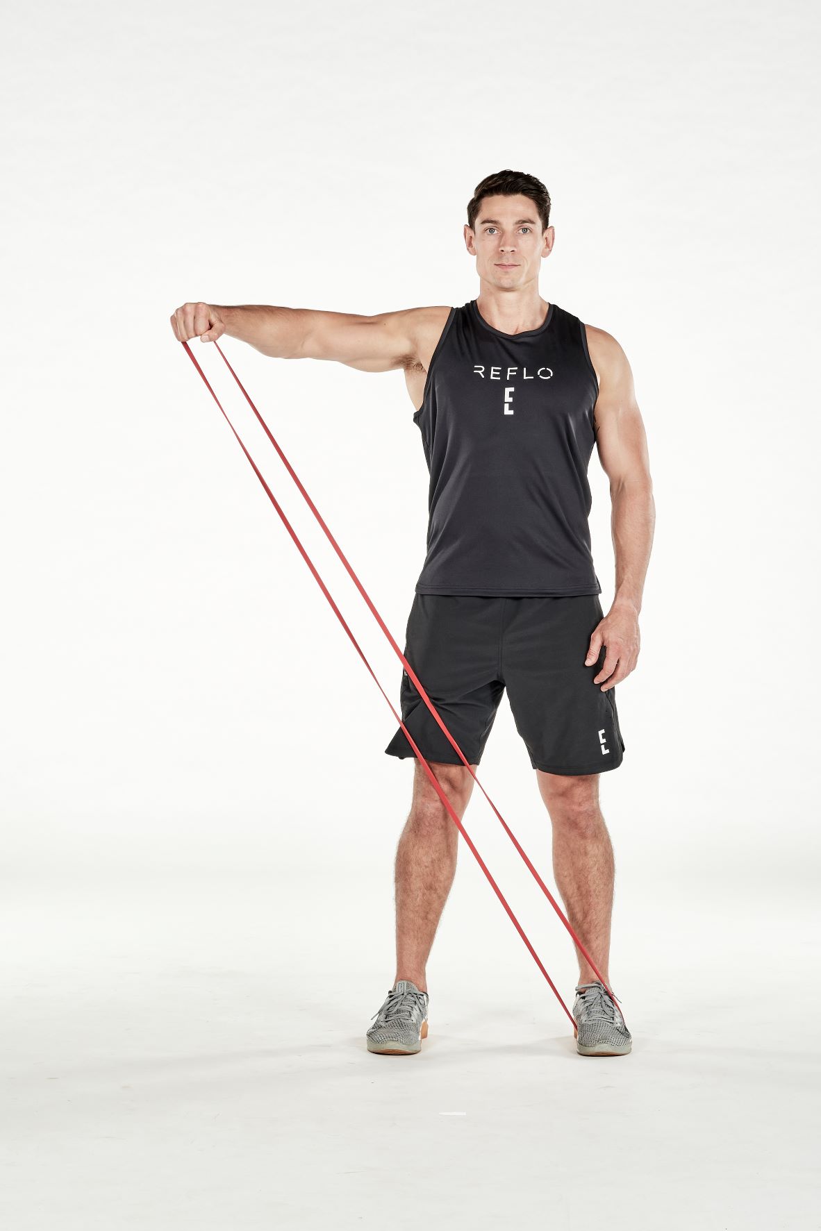 man demonstrating step two of band lateral raise; standing up straight, a band is held under one foot; holding the band with the opposite hand, he extends his arm straight out to the side until it reaches shoulder height; he wears a black fitness vest, black shorts and trainers