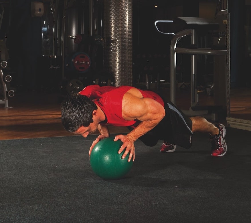 man in red vest and shorts performing the second stage of a medicine ball press-up in the gym