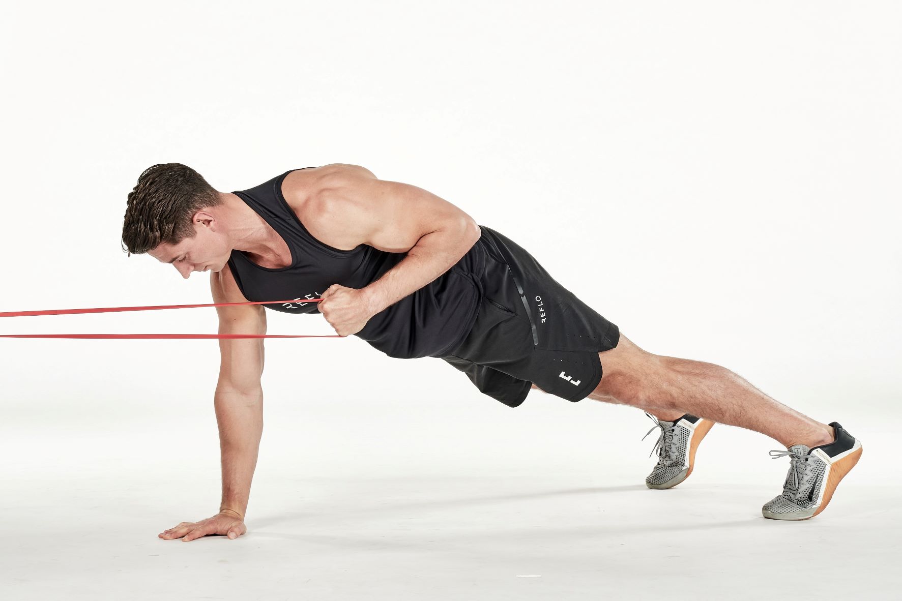 man demonstrating step two of plank row; in a plank position, one hand holds a secured band; he pulls the band in towards his chest; he wears a black fitness vest, black shorts and trainers