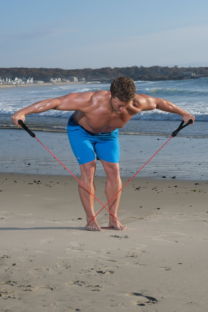 with the resistance band secured under his feet, man performs reverse flye, lifting his arms out to the side while bent forward at the hips