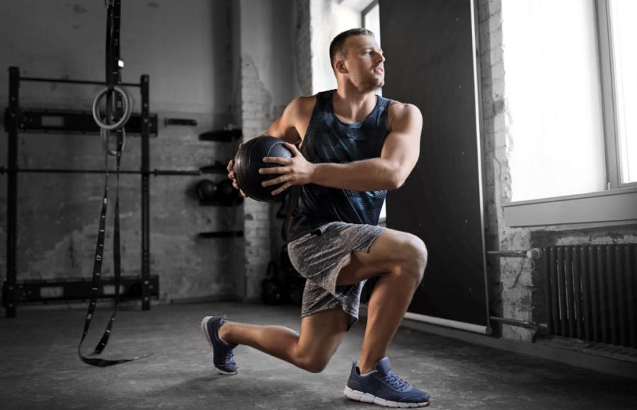 man working out with medicine ball in gym wearing gym shorts and vest top