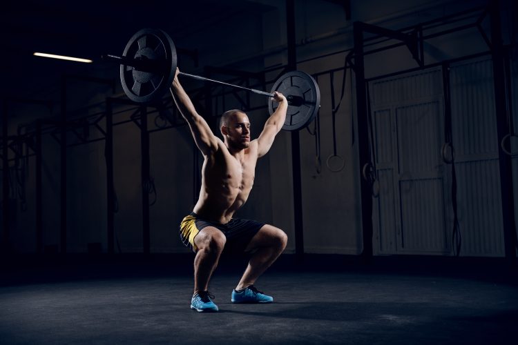 Man with bare chest squatting to lift a barbell in a gym - get stronger by training less