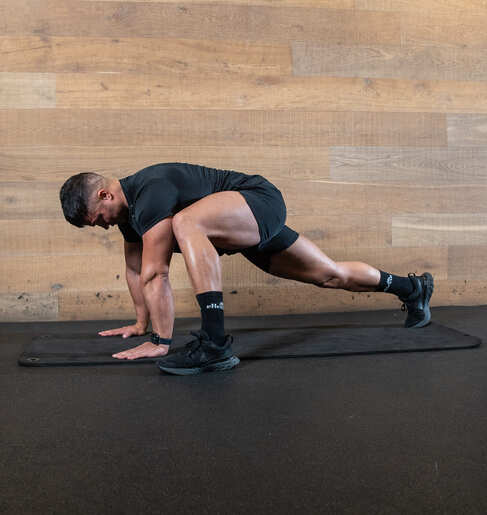 Part of the best full-body dumbbell workout: man demonstrates world's greatest stretch; from a high plank, his left legs moves to the outside of his hands